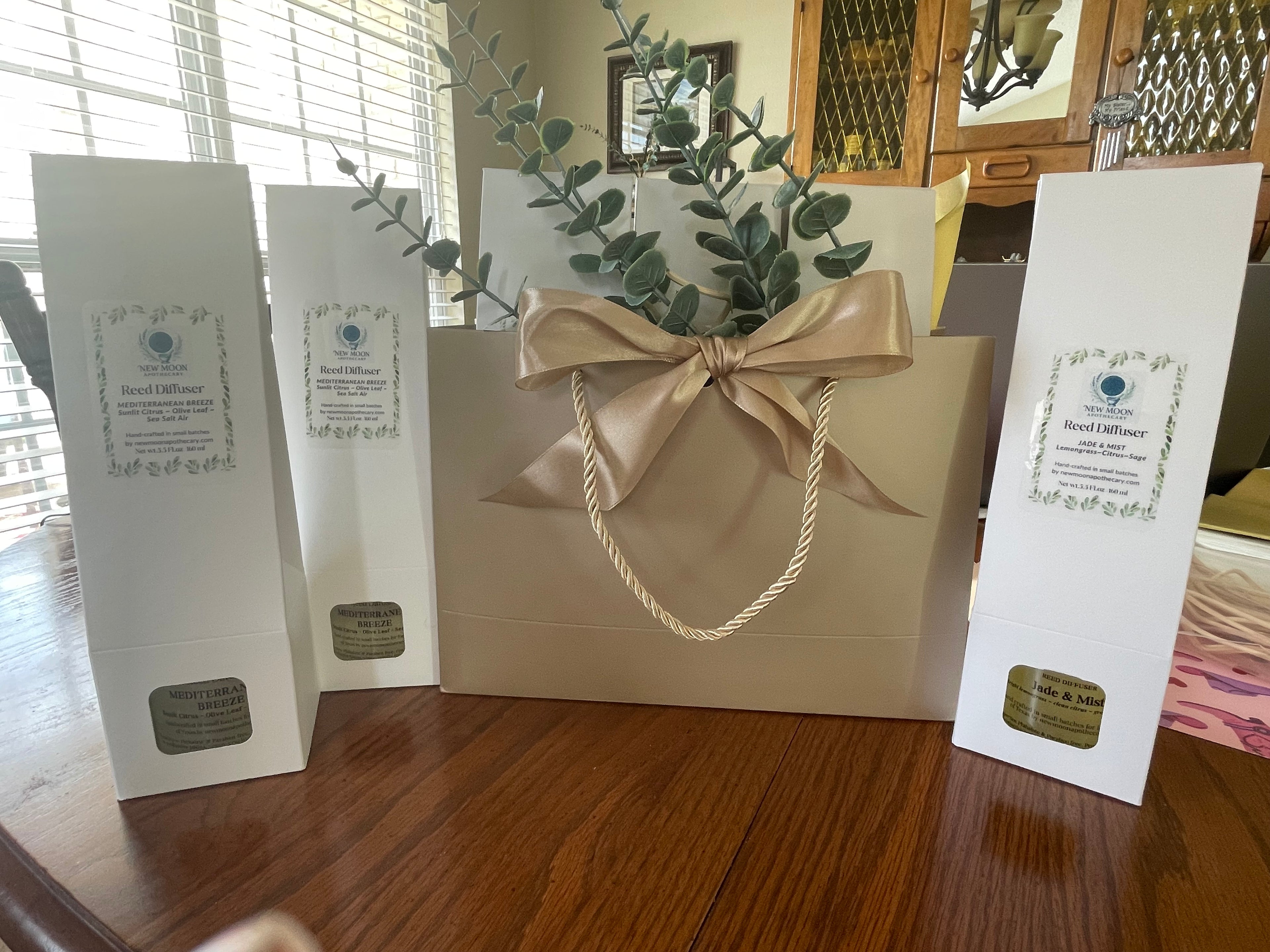 Gift bag with a bow and eucalyptus leaves, surrounded by branded boxes on a wooden surface.