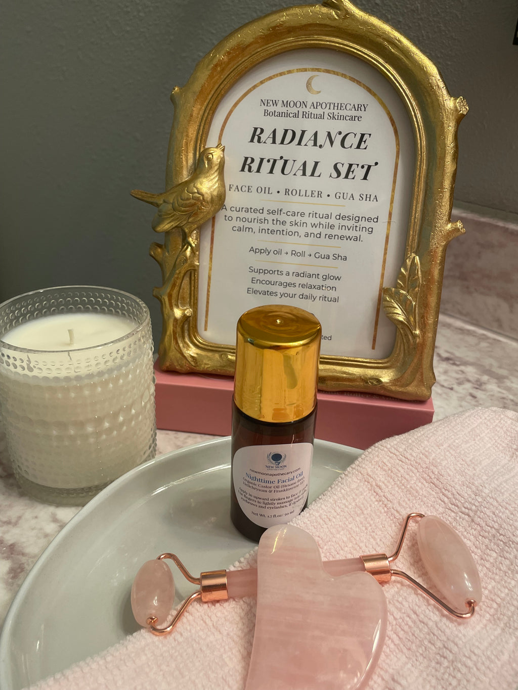 Skincare products including a bottle, roller,  gua sha and framed text on a surface. Spa candle and towel add decor to the products