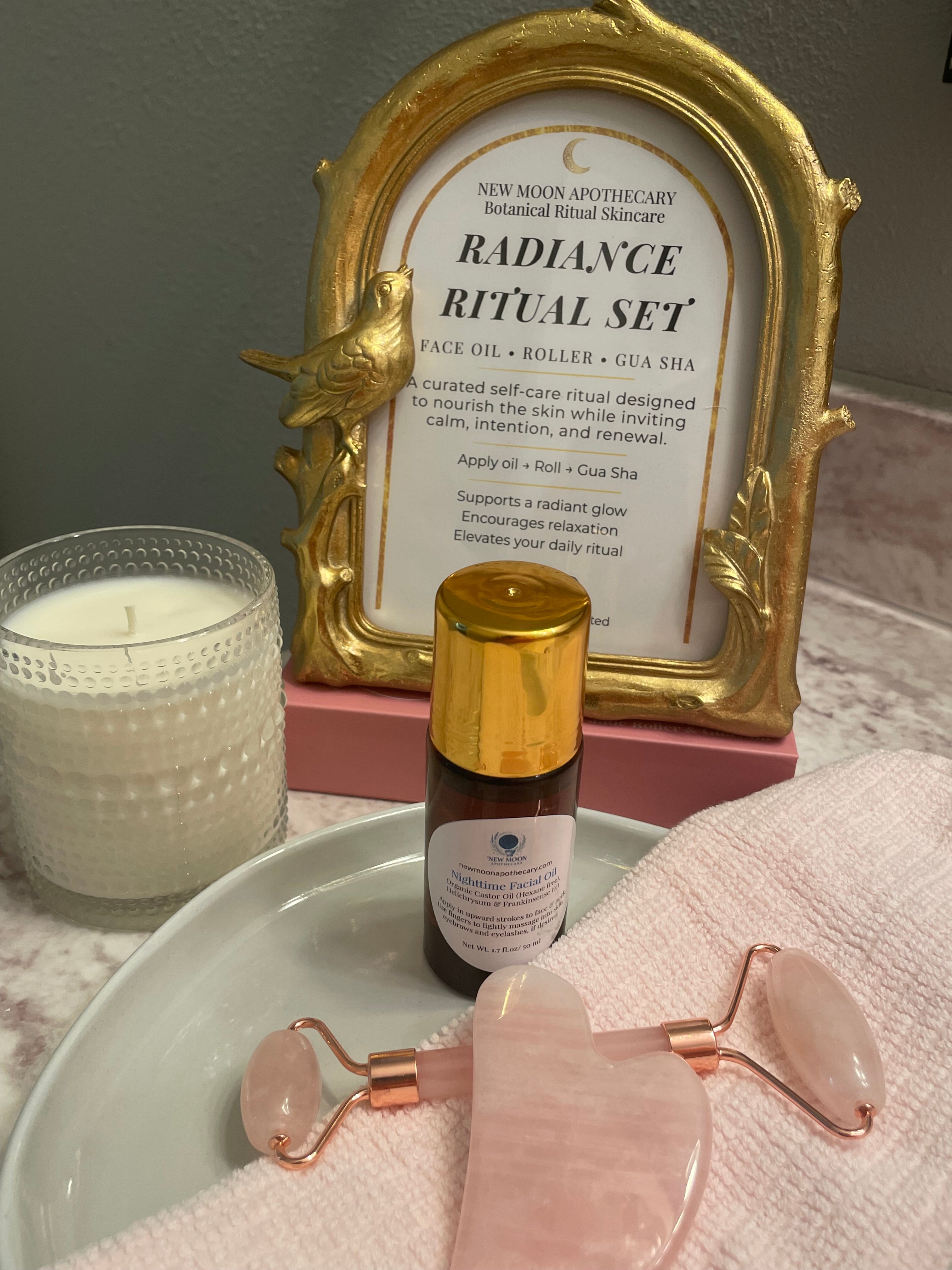 Skincare products including a bottle, roller,  gua sha and framed text on a surface. Spa candle and towel add decor to the products