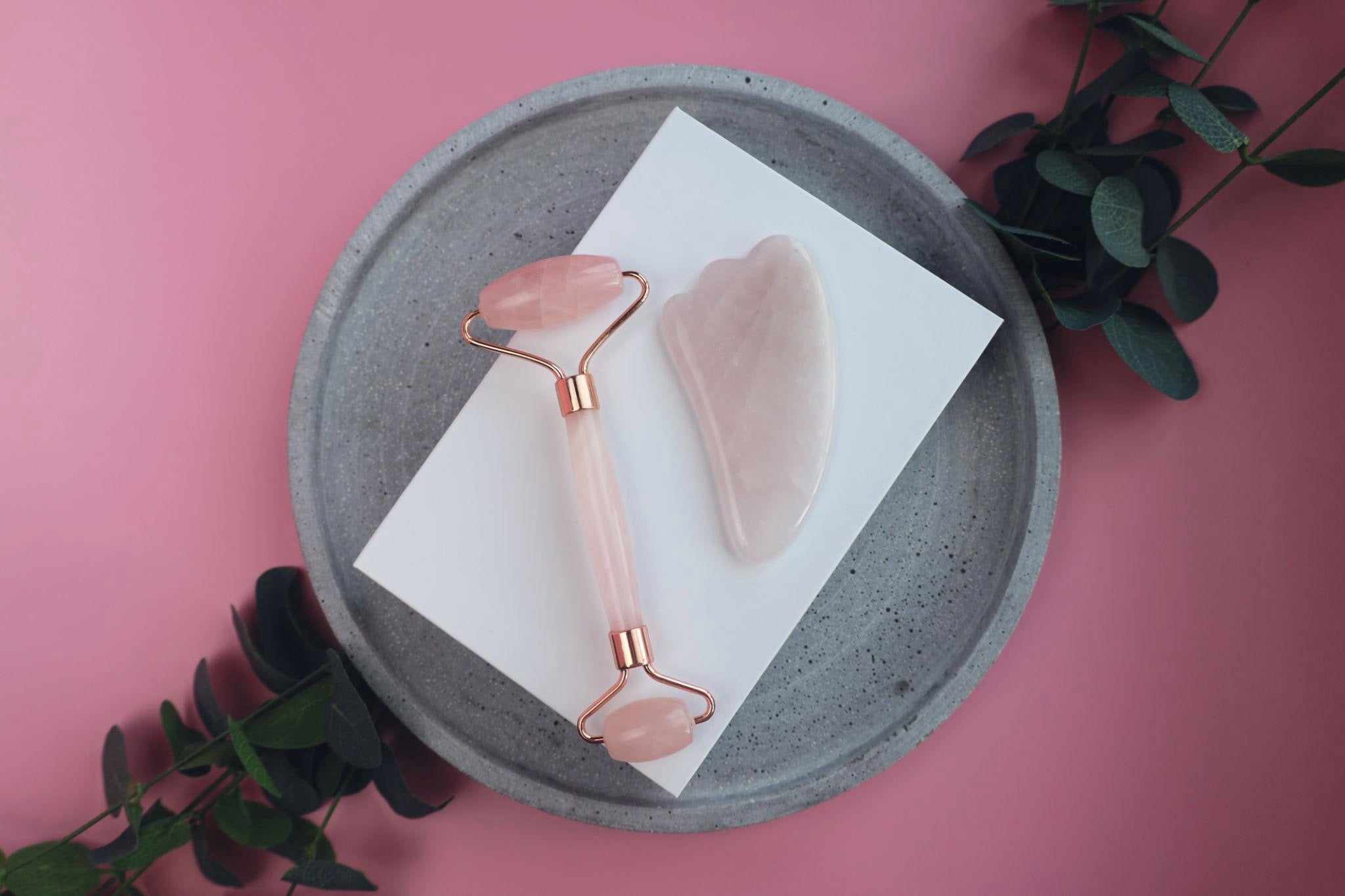 Rose quartz roller and gua sha tool on a gray plate with a pink background